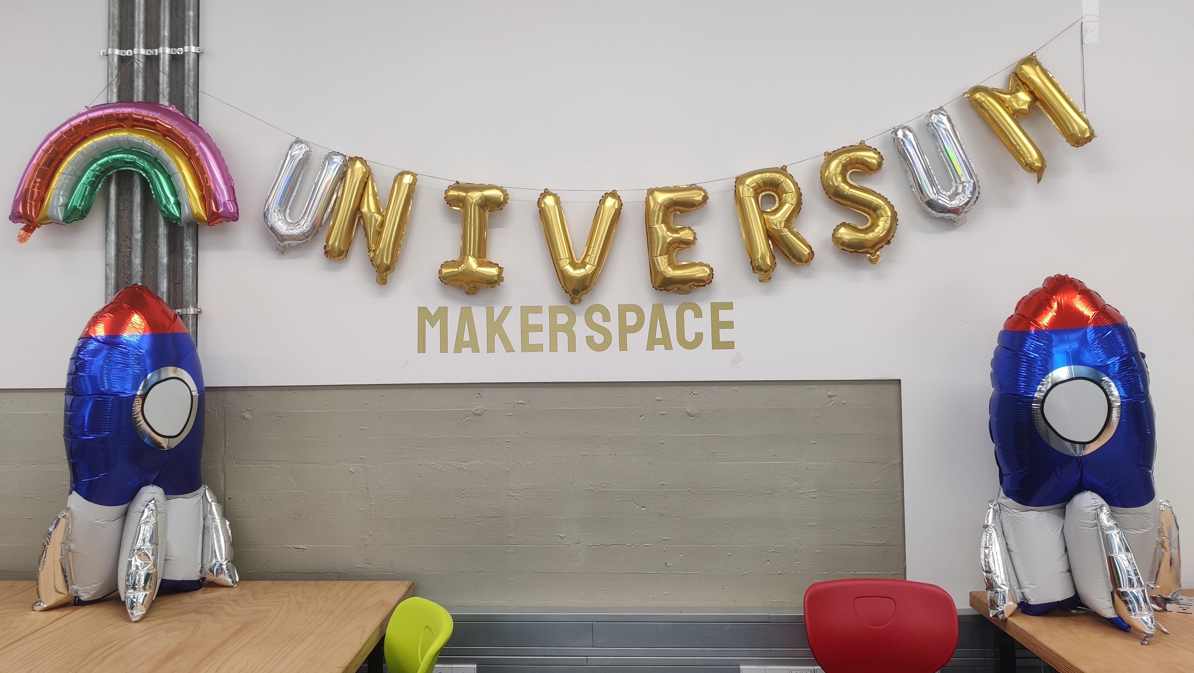 Luftballons an der Wand im Makerspace bilden den Schriftzug "Universum", darunter steht als Wandsticker "Makerspace", eingerahmt ist das ganze von zwei Raketen-Ballons und einem Regenbogen-Ballon.